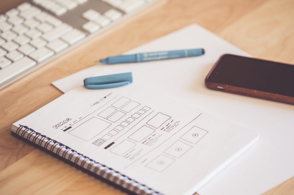 A neat workspace featuring a sketchpad with wireframes, a smartphone, and a keyboard on a wooden desk.